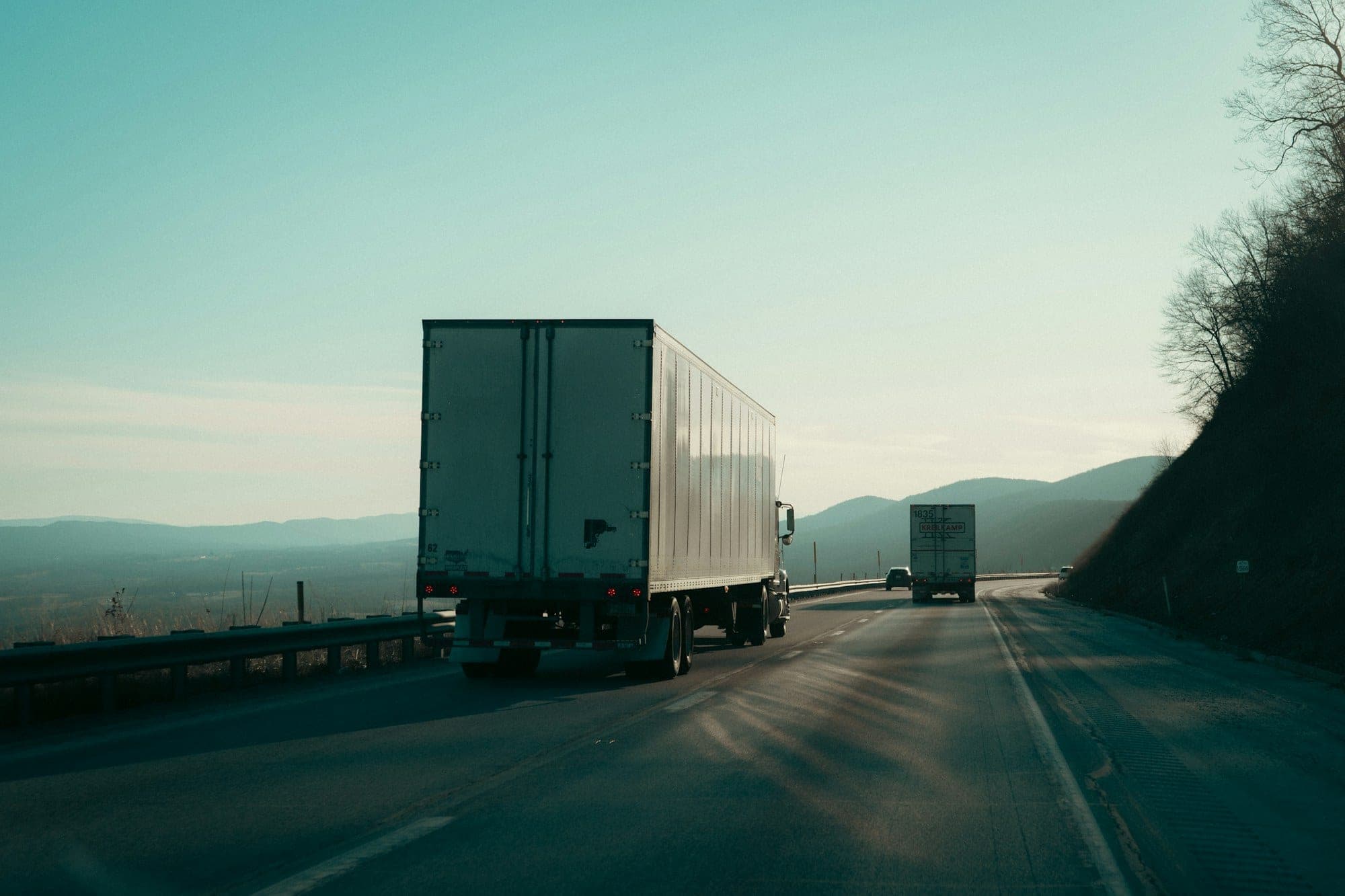 Driver's-eye view of two semi trucks with dry van trailers climbing a mountain interstate in low afternoon sun