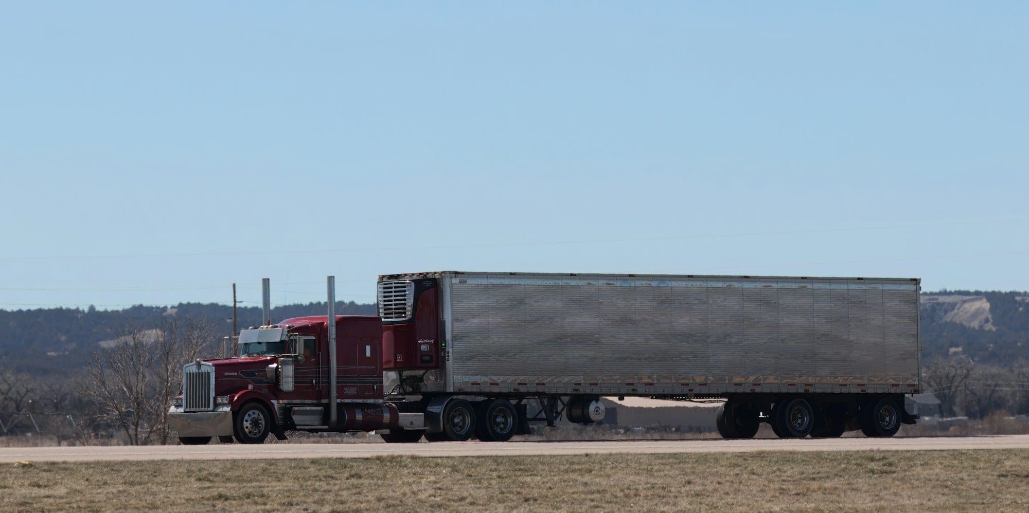 Side view of a maroon long-nose semi truck pulling a silver refrigerated trailer along Interstate 80 in Nebraska