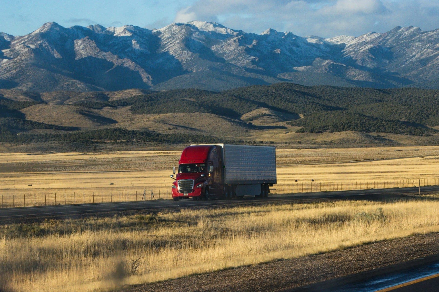 Red semi truck pulling a silver dry van trailer through golden grassland beneath snow-capped mountains