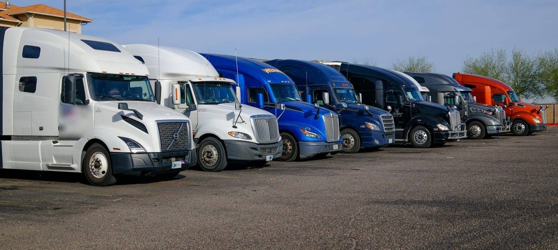 Row of modern semi truck tractors in white, blue, black and orange parked side by side under a blue sky