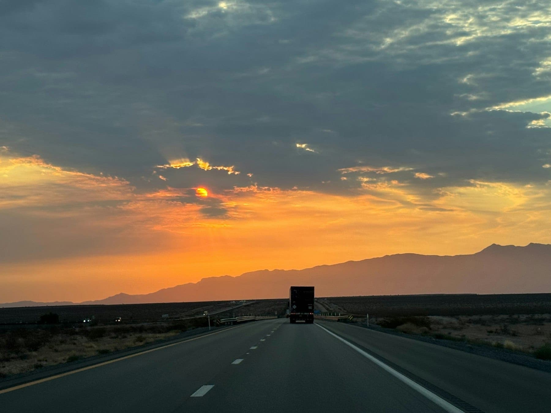 Semi truck driving down a long, straight interstate toward a mountain range under a dramatic orange sunset sky