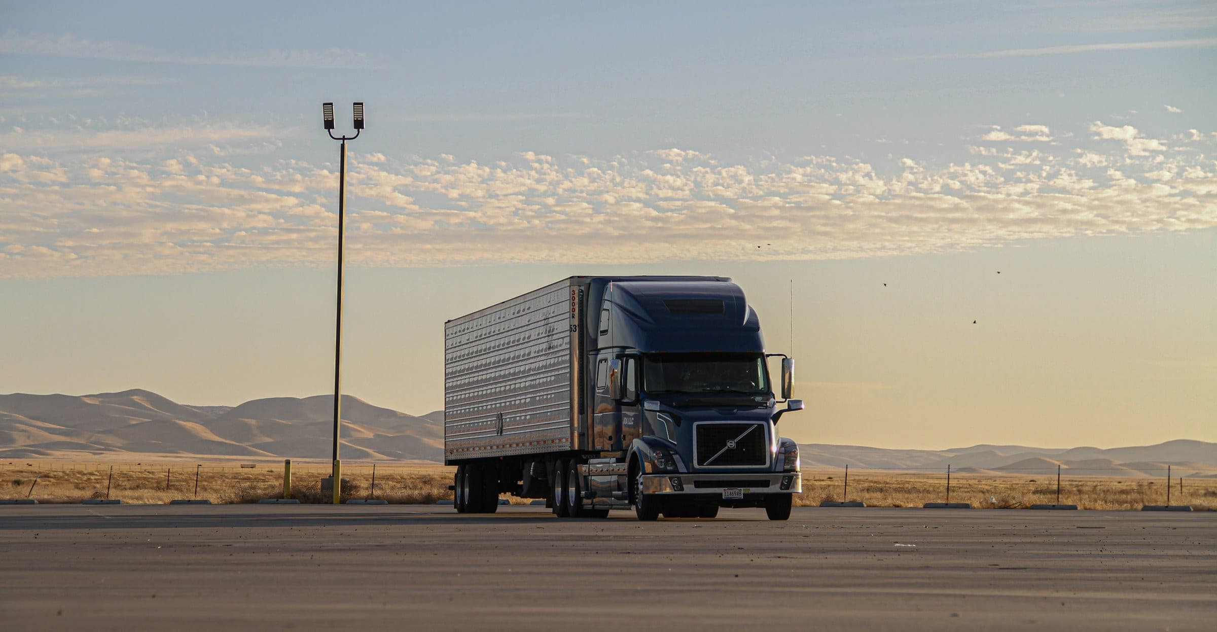 Dark blue Volvo semi truck with a refrigerated trailer under a warm evening sky, with golden hills on the horizon