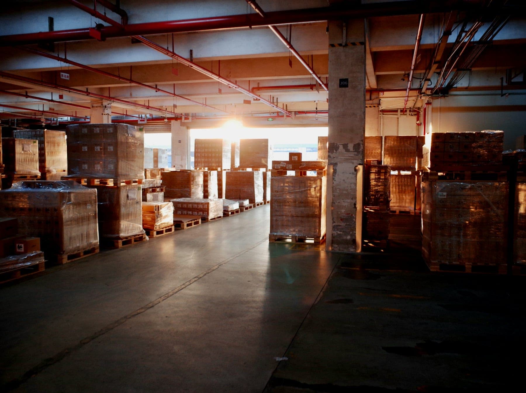 Warehouse floor lined with shrink-wrapped pallets of goods as sunlight streams through an open dock
